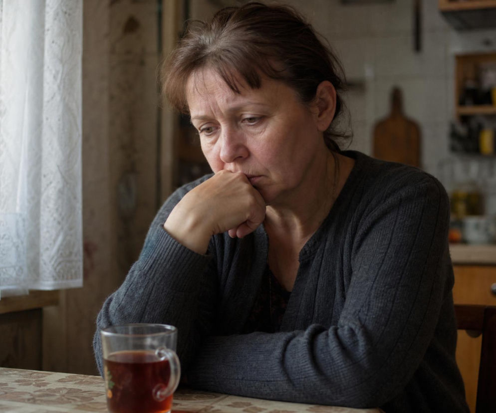 Zmartwiona kobieta w średnim wieku siedzi przy stole z kubkiem herbaty, patrząc w dół z ręką na ustach, wyrażając smutek i rozczarowanie. Scena z artykułu Przeniosłam pieniądze Zosi na lepszą lokatę na naszym portalu.