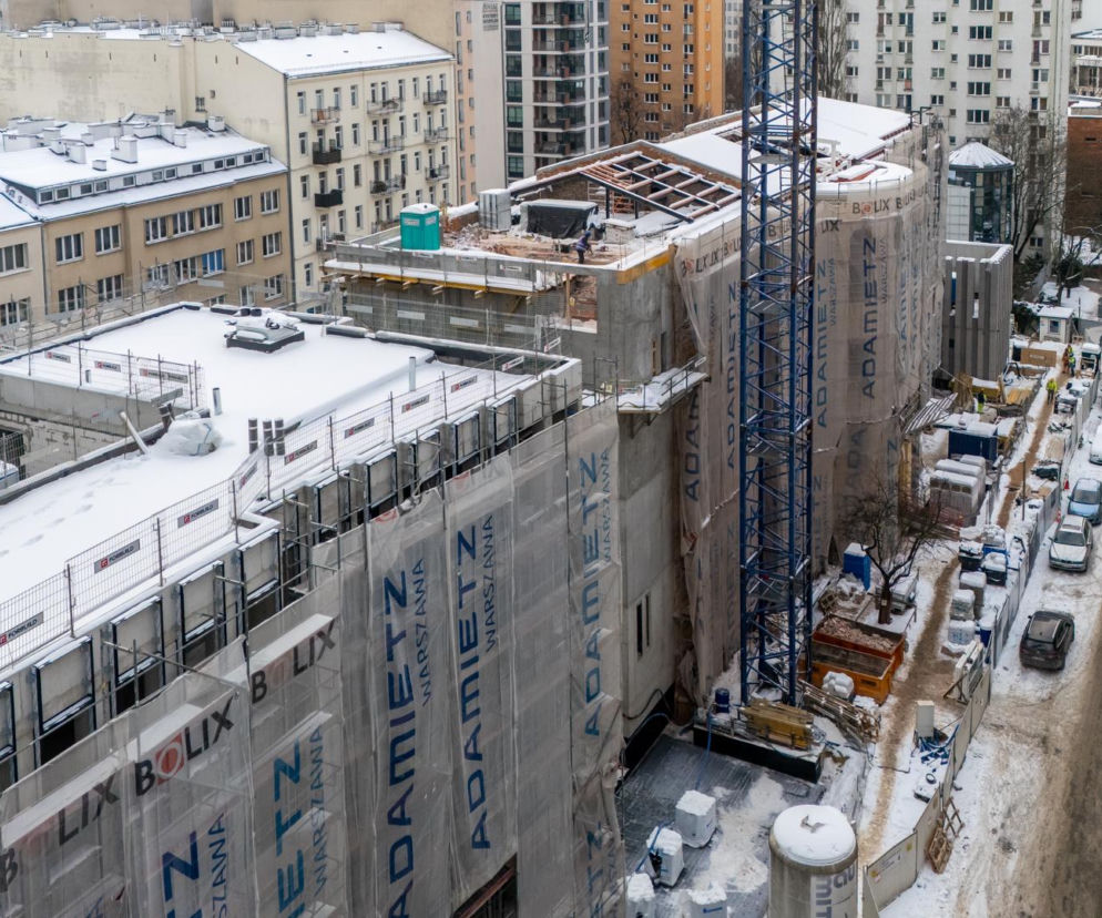 Zimowy widok z góry na plac budowy Muzeum Getta Warszawskiego, gdzie trwają prace wykończeniowe. Nowa konstrukcja, częściowo osłonięta siatkami z logo Adamietz, pokryta jest śniegiem, podobnie jak dachy okolicznych kamienic. Artykuł o postępach budowy na Architektura Murator Plus.
