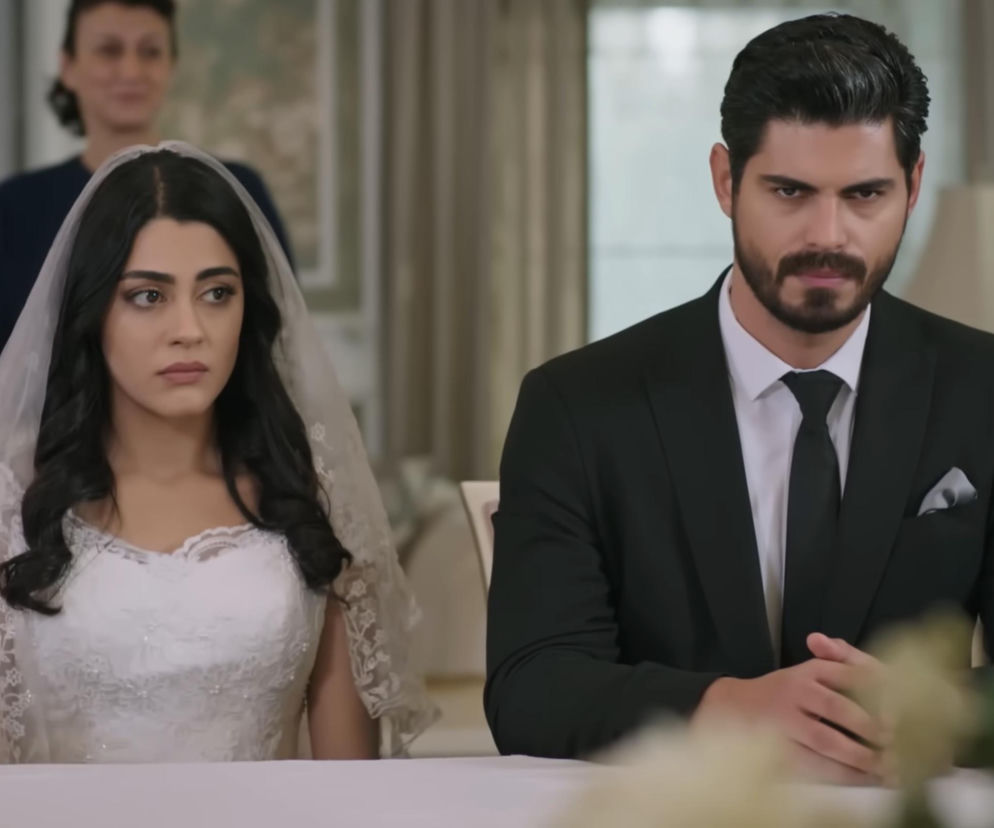 Panna młoda Hancer w białej sukni ślubnej i welonie, z czarnymi włosami i smutnym wyrazem twarzy, siedzi obok Cihana w garniturze z kamienną miną. Za nimi stoi kobieta. Smutny ślub Hancer i Cihana z serialu Panna młoda i inne wiadomości znajdziesz na Super Seriale.