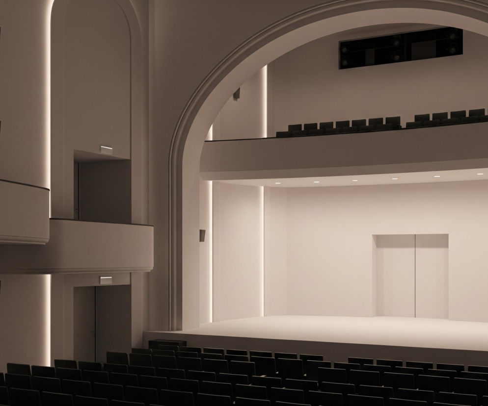 Wnętrze teatru lub sali koncertowej o stonowanej kolorystyce z dominującymi odcieniami beżu i bieli. Na pierwszym planie widoczne są rzędy ciemnych, pustych foteli, skierowanych w stronę podwyższonej sceny w kolorze écru, która znajduje się na drugim planie. Ściana za sceną jest również jasna, z dużym, dwuskrzydłowym otworem drzwiowym po prawej stronie. Po bokach sceny biegną pionowe pasy podświetlenia LED, a na lewej ścianie, poza głównym obszarem sceny, znajdują się dwa balkony w kształcie półokręgu, z czarnymi poręczami i wnękami podświetlonymi podobnymi do tych na scenie. Nad sceną, w górnej części, widać drugi rząd pustych foteli na balkonie, a powyżej niego, w dużej wnęce, umieszczono czarne elementy nagłośnienia lub oświetlenia, wszystko to w obrębie dużego, łukowatego przejścia.