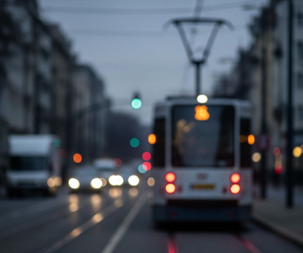 Na pierwszym planie, centralnie, widoczny jest tył białego tramwaju z dwoma czerwonymi, okrągłymi światłami hamowania oraz pomarańczowym wyświetlaczem z cyframi u góry. Nad tramwajem znajduje się rozmyty pantograf i jasne, małe światło. Ulica przed tramwajem jest ciemna, z odbiciami czerwonych świateł na torach. Po lewej stronie w oddali jadą inne rozmyte pojazdy, w tym biały samochód osobowy i biała ciężarówka, których światła przednie świecą na jasnożółto. Tło stanowią rozmyte budynki po obu stronach ulicy, a w oddali widać kolorowe, rozmyte światła, w tym zielone, pomarańczowe i czerwone punkty, sugerujące światła drogowe lub inne źródła światła miejskiego.
