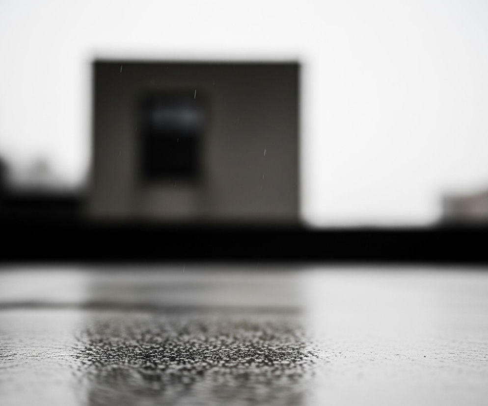 Pierwszy plan ukazuje mokrą, ciemną powierzchnię, pokrytą licznymi kropelkami wody, które odbijają światło, tworząc efekt iskrzenia. Powierzchnia ta rozciąga się od dołu obrazu, lekko wznosząc się ku górze, a jej faktura staje się mniej wyraźna w oddali. Na drugim planie, poza ostrością, widoczny jest szarobury budynek o prostokątnym kształcie z ciemnym, centralnie umieszczonym otworem przypominającym okno lub wejście. W powietrzu, zwłaszcza w okolicach budynku, można dostrzec pojedyncze, cienkie smugi, sugerujące padający deszcz, a całe tło jest jasne i rozmyte, co wskazuje na pochmurny dzień.