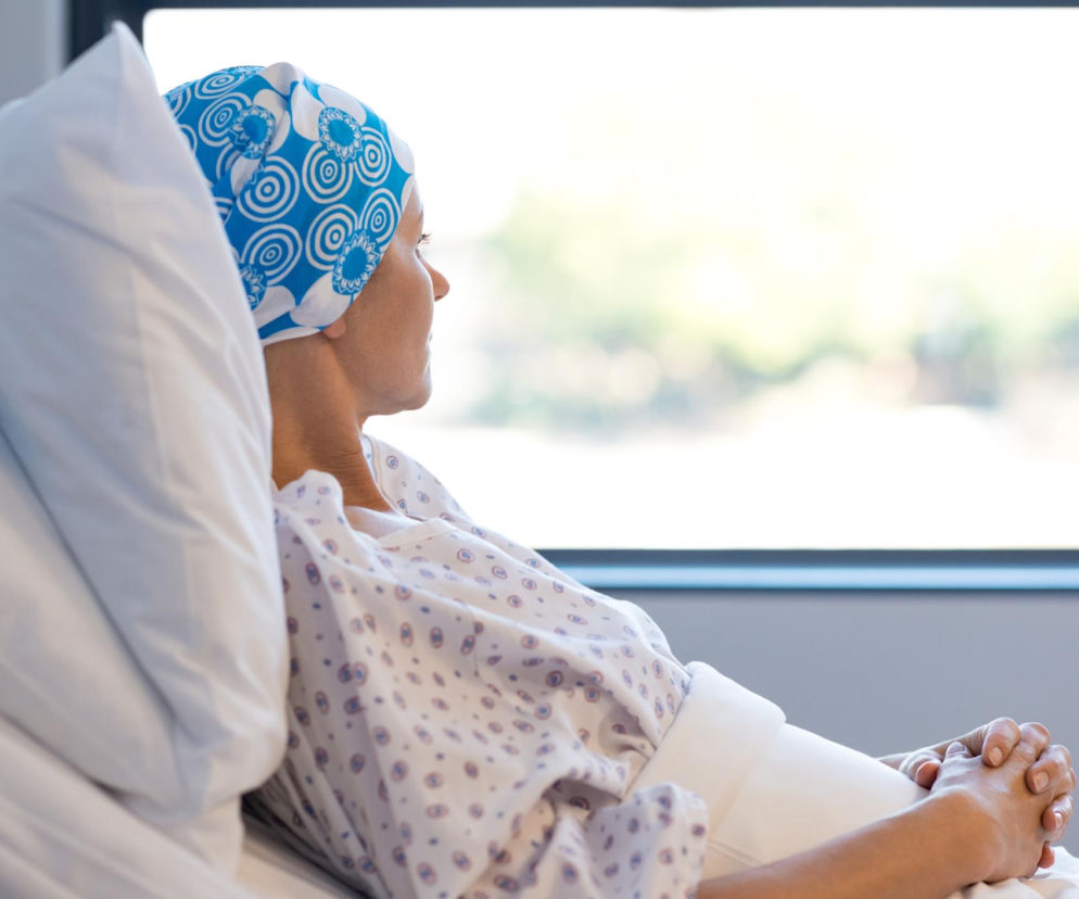 Kobieta w niebieskiej chustce na głowie i szpitalnej koszuli siedzi na łóżku, spoglądając przez okno. Złączone dłonie symbolizują nadzieję i oczekiwanie na pozytywne efekty leczenia glejaka, o czym przeczytasz na Poradnik Zdrowie.