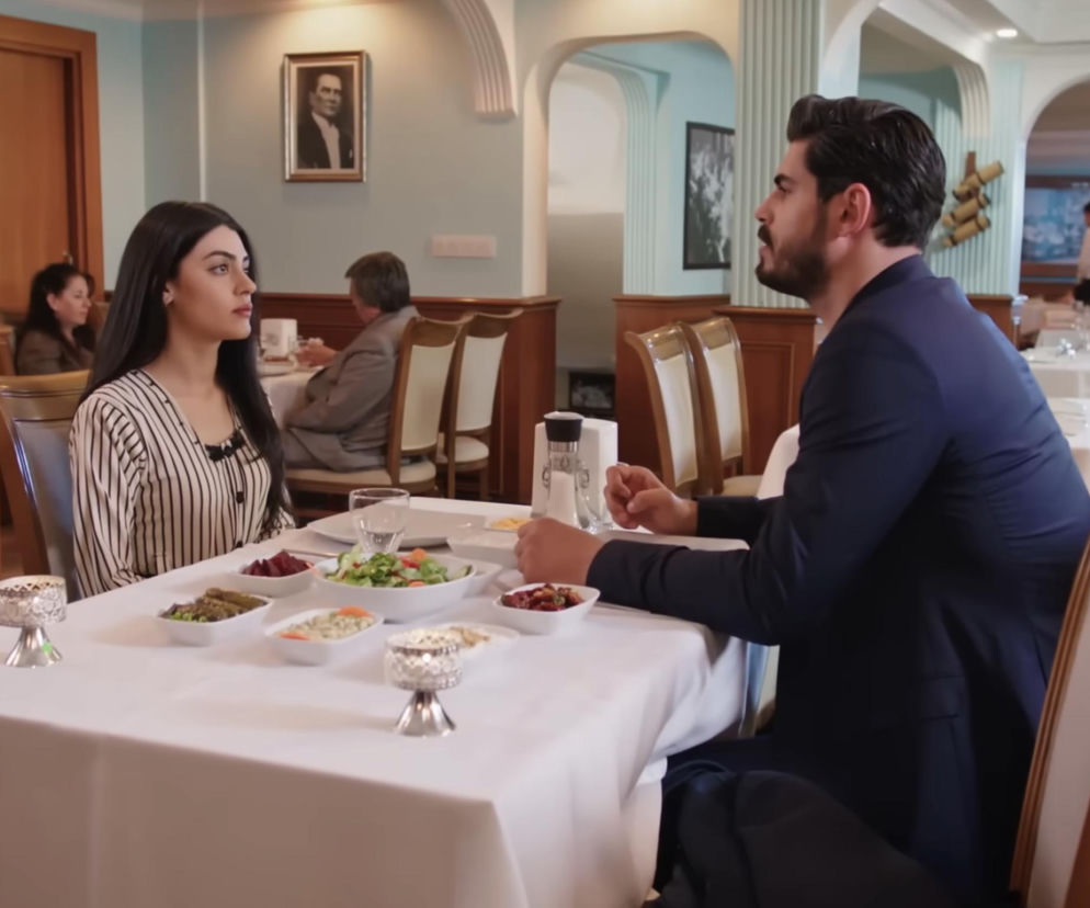 Hancer i Cihan siedzą przy stole w restauracji, próbując odbudować relacje. Hancer, ubrana w pasiastą bluzkę, unika wzroku Cihana, który w granatowej marynarce próbuje zamówić jedzenie. Scena z serialu Panna Młoda dostępna na Super Seriale.
