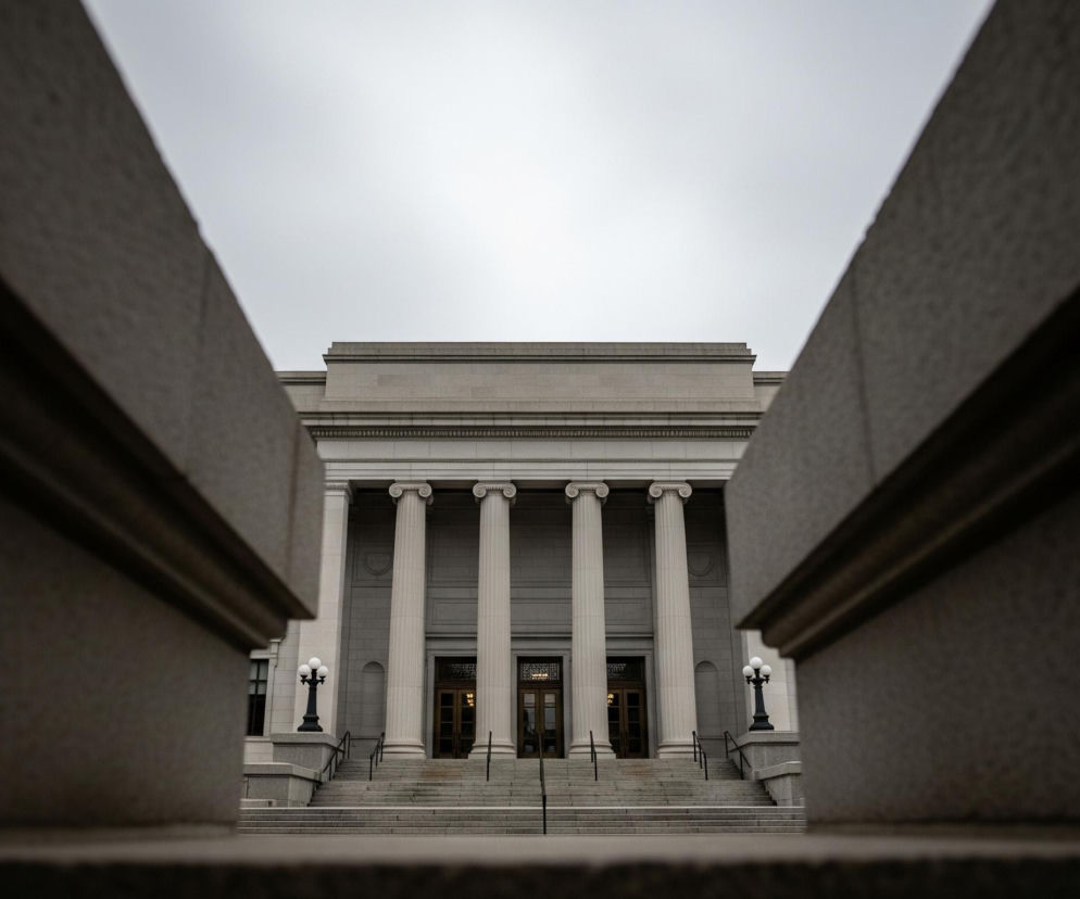 Centralnym punktem obrazu jest duży, monumentalny budynek z jasnego kamienia, ujęty w kadrze z perspektywy z dołu. Fasada budynku charakteryzuje się rzędem sześciu wysokich, rzeźbionych kolumn z jońskimi kapitelami, wspierających prosty gzyms i attykę. Poniżej kolumn znajdują się trzy ciemne, prostokątne wejścia z przeszklonymi drzwiami, do których prowadzą szerokie, kamienne schody z ciemnymi, metalowymi balustradami. Po obu stronach schodów stoją czarne latarnie z trzema kulistymi kloszami, a po bokach kadru widoczne są fragmenty ciemnych, kamiennych balustrad, które tworzą swoistą ramę dla budynku. Jasne, jednolite niebo zajmuje górną część obrazu.