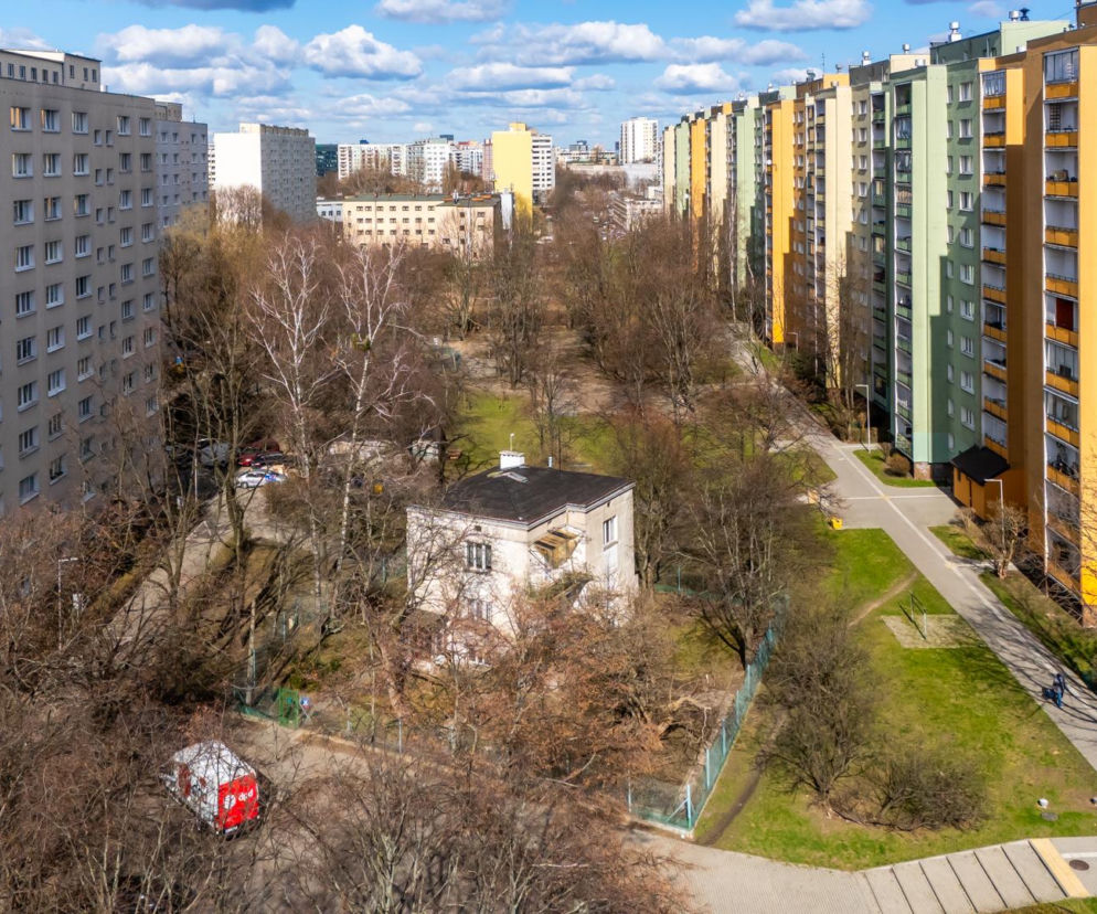 Murowany piętrowy dom jednorodzinny stoi pod skosem między wysokimi blokami. O historii budynku przeczytasz na Architektura.