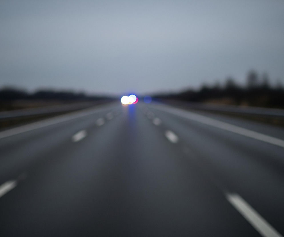 Droga asfaltowa, z białymi, przerywanymi liniami pasa ruchu, rozciąga się w głąb obrazu. W oddali, na środku drogi, widoczne są rozmyte, intensywnie świecące punkty świetlne: dwa białe, jeden czerwony i jeden niebieski, sugerujące światła pojazdu. Po obu stronach drogi rozciąga się zamazany, ciemny pas, przypominający zalesiony teren lub krzewy, nad którym unosi się jasnoszare, pochmurne niebo.