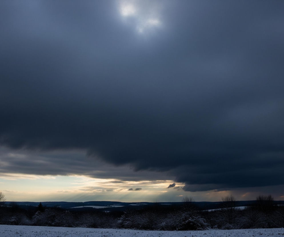 Na zdjęciu dominuje ciemne, zachmurzone niebo w odcieniach szarości i granatu, z jaśniejszymi smugami chmur w górnej części, skąd przebija się słońce, tworząc promieniste rozbłyski. Poniżej, w środkowej części, horyzont rozświetlają złoto-pomarańczowe i jasnoniebieskie odcienie, z których widoczne są wyraźne promienie światła padające na odległe, pofalowane wzgórza i pola. Pierwszy plan zajmuje zaśnieżony, biały krajobraz z ciemnymi sylwetkami drzew i krzewów, pokrytych delikatną warstwą śniegu.