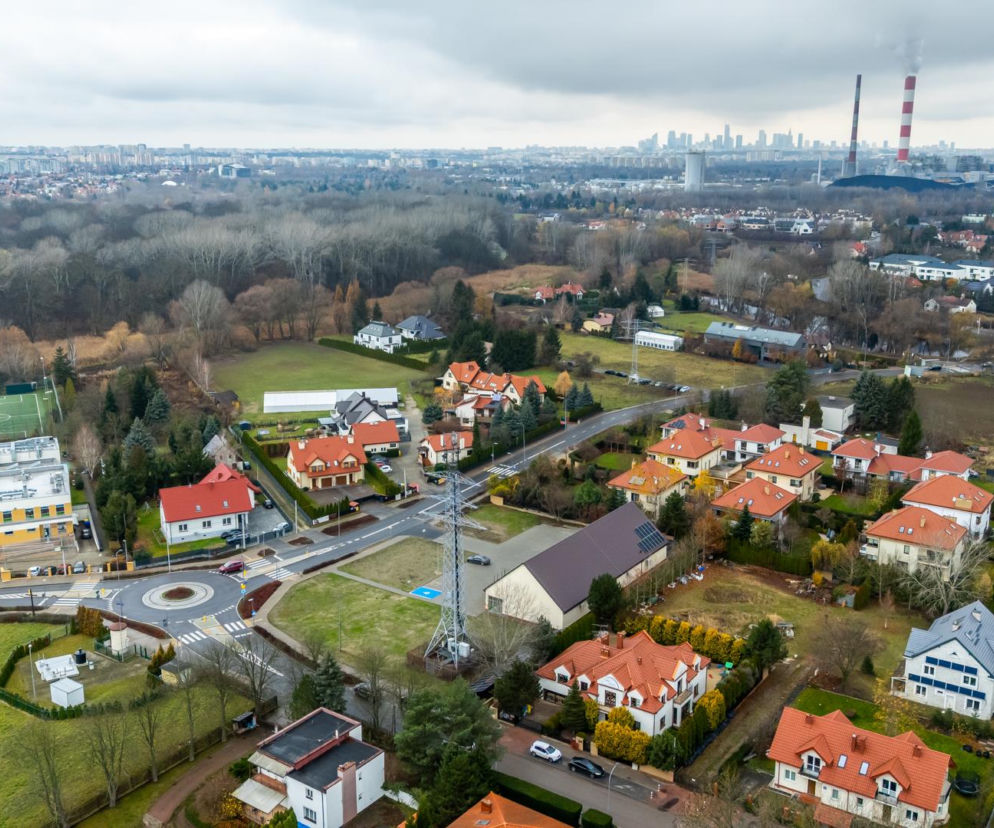 Widok z lotu ptaka na Wilanów Zawady z zabudową jednorodzinną i lasem, w tle wieżowce Warszawy i kominy elektrociepłowni, co symbolizuje wyzwania planistyczne. O konsekwencjach paraliżu budowlanego po 2026 roku przeczytasz na Super Biznes.