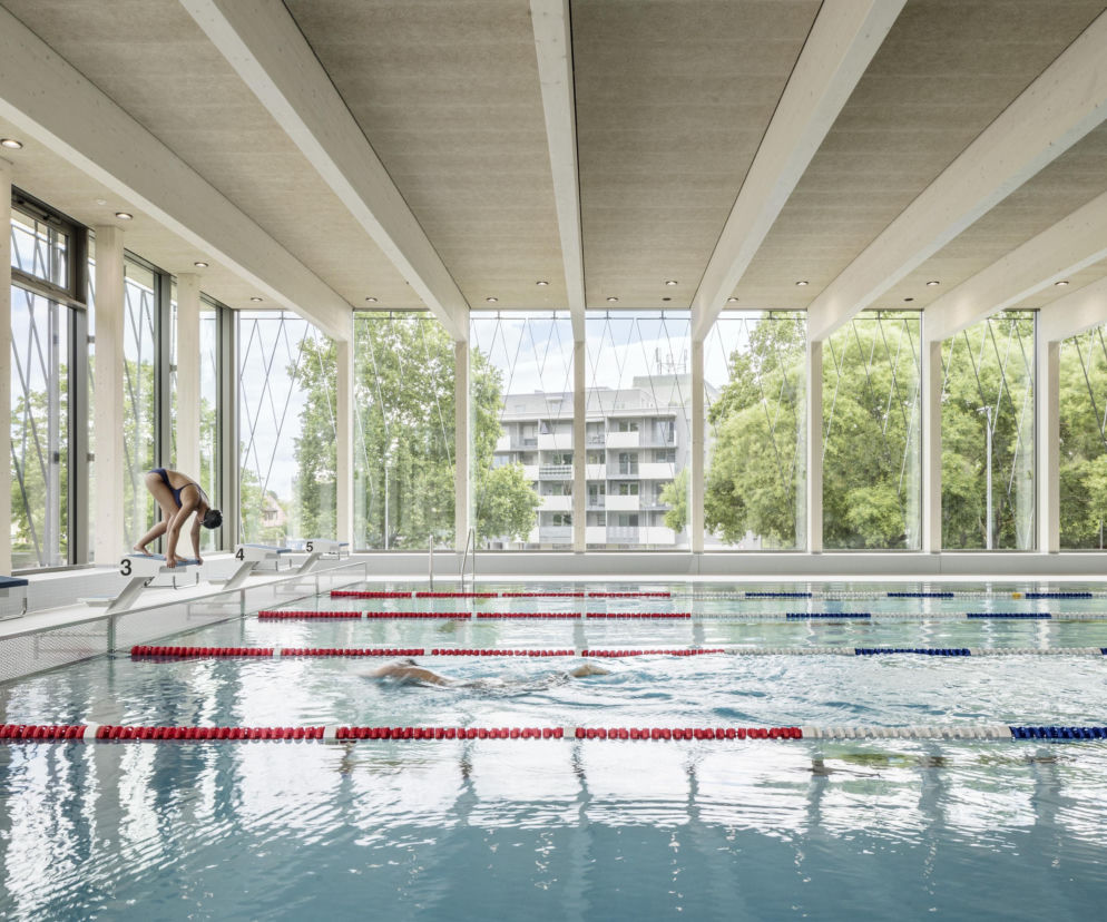Nowoczesny basen z dużymi oknami, przez które widać zieleń i budynki mieszkalne. Pływaczka przygotowująca się do startu na torze 3 oraz osoby w wodzie. O architekturze takich obiektów przeczytasz na portalu Architektura Murator Plus.