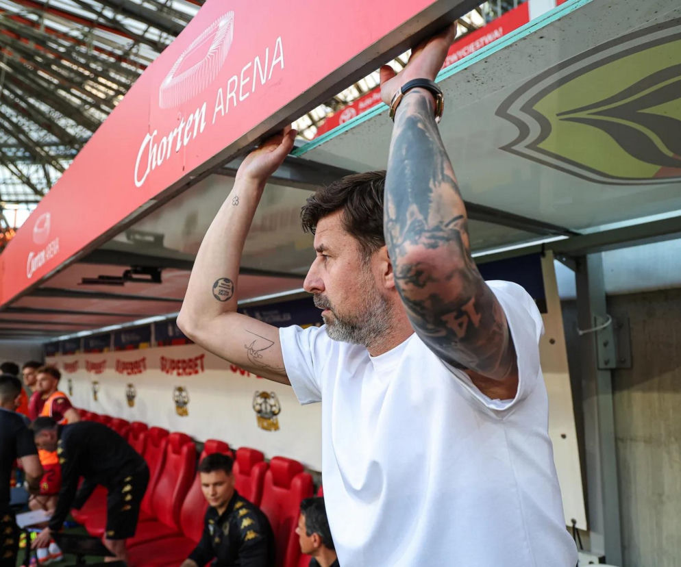 Łukasz Masłowski, menedżer z tatuażami na ramionach, stoi w białej koszulce na tle stadionu Chorten Arena z widocznymi na trybunach kibicami, opierając się o bandę. Artykuł o jego nowym klubie przeczytasz na naszym portalu.