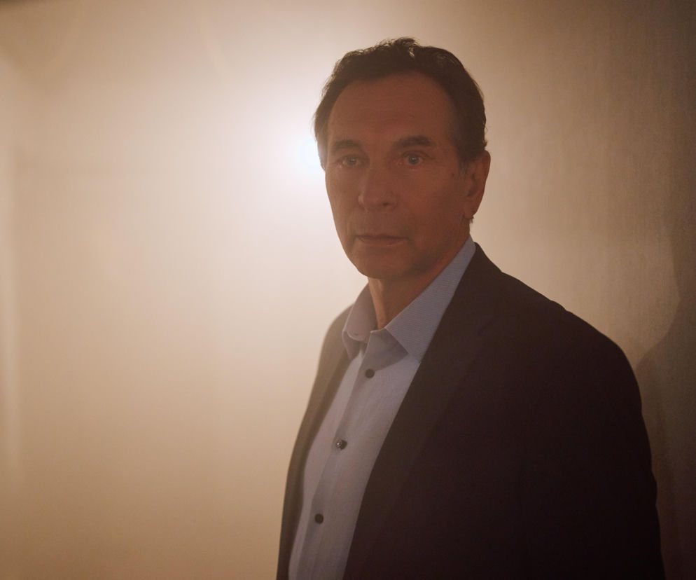 Tomasz Urbanek, aktor znany z roli w serialu Na Wspólnej, ubrany w elegancką marynarkę i jasną koszulę, patrzy prosto w obiektyw, stojąc na tle delikatnie oświetlonej ściany. Zdjęcie w kontekście dramatycznych wydarzeń w pensjonacie Hofferów znajdziesz na Super Seriale.