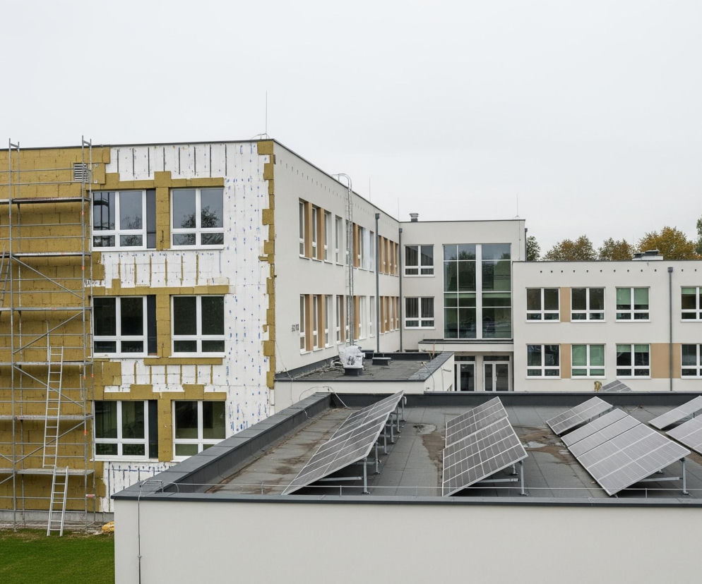 Budynek w trakcie termomodernizacji, ze ścianą szczytową pokrytą żółtą wełną mineralną i częściowo białą folią wiatroizolacyjną. Z boku tej ściany znajduje się rusztowanie z metalowymi drabinkami, a na ścianach fasadowych widoczne są liczne okna z białymi ramami. Na płaskim dachu przed budynkiem rozmieszczono panele fotowoltaiczne w kilku rzędach, z widocznymi kałużami wody na papie. W tle widoczne są drzewa z zielonymi i pomarańczowymi liśćmi pod pochmurnym, jasnoszarym niebem.