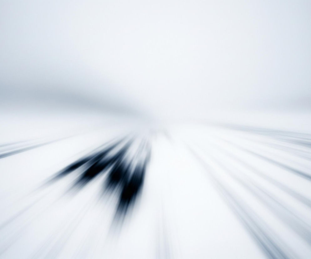 Rozmyte, dynamiczne ujęcie w odcieniach bieli i błękitu. Centralnie, w dolnej połowie kadru, widoczne są trzy niewyraźne, ciemne sylwetki o wydłużonym kształcie, sugerujące ruch w stronę obserwatora. Od tych sylwetek oraz z dolnych krawędzi kadru rozchodzą się promieniście liczne, jasne i ciemne smugi, akcentujące wrażenie szybkości i ruchu. Górna część obrazu to jaśniejsze, niemal białe tło, które stopniowo ciemnieje w stronę dolnych partii, zlewając się z rozmyciami.