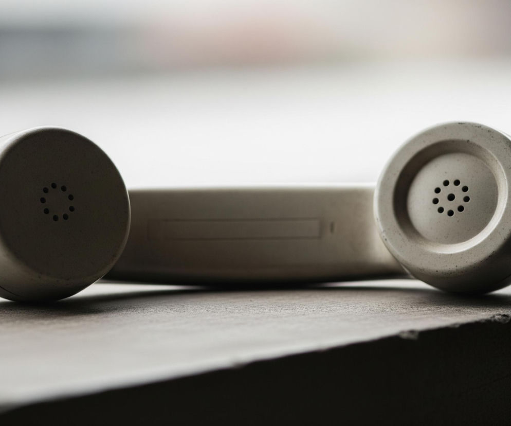 Na drewnianej lub ciemnej powierzchni leży beżowa lub jasnoszara słuchawka starego telefonu. Widoczne są oba jej zaokrąglone końce, z otworami pośrodku każdego, sugerującymi mikrofon i głośnik. Między nimi znajduje się prostokątny korpus słuchawki, a od lewego końca odchodzi spiralny kabel, który biegnie w głąb obrazu. Tło jest rozmyte i jasne, z niewielkimi odcieniami szarości i bieli, co skupia uwagę na słuchawce.