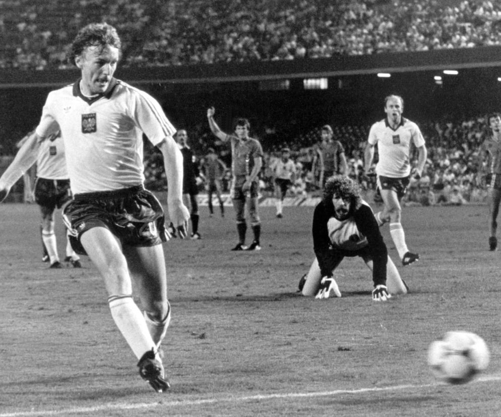 Zbigniew Boniek w białej koszulce z orzełkiem i ciemnych spodenkach, biegnący z piłką obok klęczącego bramkarza na meczu mundialu w 1982 roku. Kibice na trybunach i inni piłkarze w tle. Więcej o karierze Bońka przeczytasz na naszym portalu.