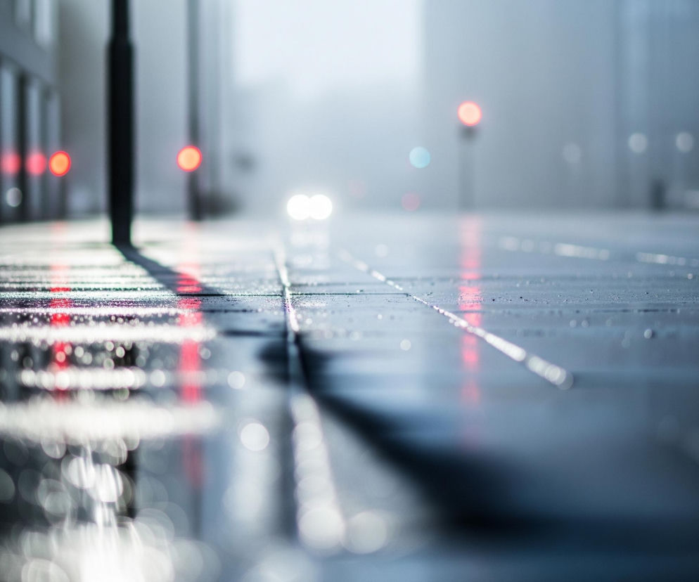 Mokra, ciemnoszara nawierzchnia drogi z widocznymi refleksami świateł zajmuje dolną połowę kadru. Światła odbijają się od powierzchni, tworząc rozmyte kręgi i smugi, z dominującymi czerwonymi i białymi barwami. W oddali, na rozmytym tle miejskim, widać dwie jasne, białe plamy świateł samochodu oraz kilka rozmazanych, czerwonych świateł, prawdopodobnie sygnalizacji lub tylnych świateł innych pojazdów. Po lewej stronie wznosi się ciemny, pionowy słup, rzucający długi cień na mokrą drogę, a za nim widać rozmazane kształty budynków.