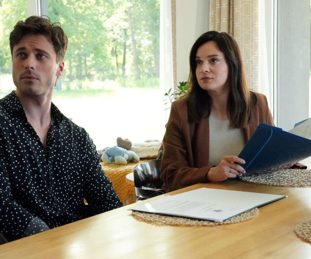 Natalia (Maria Dejmek) i Cezary (Marcel Opaliński) siedzą przy drewnianym stole w jasnym wnętrzu. Natalia trzyma niebieską teczkę z dokumentami, a Cezary patrzy w bok z zamyśloną miną, co wskazuje na kryzys w ich związku. Więcej o ich losach w Super Seriale.