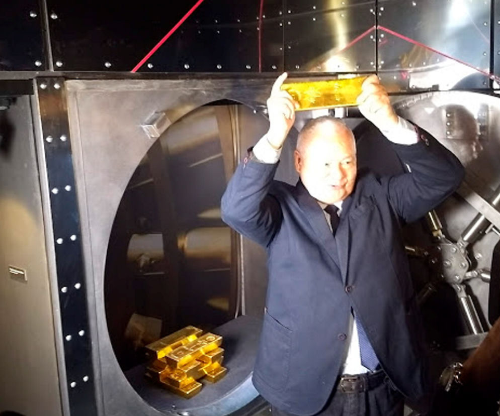 Prezes NBP Adam Glapiński, w marynarce i koszuli, unosi sztabkę złota przed otwartym sejfem pełnym złota. Tło to metalowe ściany z czerwoną linią LED. Więcej o kontroli NIK w NBP przeczytasz na portalu Super Biznes.