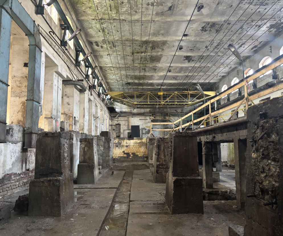Wnętrze opuszczonej hali Królewskiej Papierni w Konstancinie-Jeziornie, z widocznymi zniszczonymi elementami konstrukcyjnymi i śladami po demontażu maszyn. Betonowy sufit z widocznymi instalacjami i liczne słupy, tworzące przestrzeń wymagającą rewitalizacji, o której przeczytasz na Architektura Murator Plus.