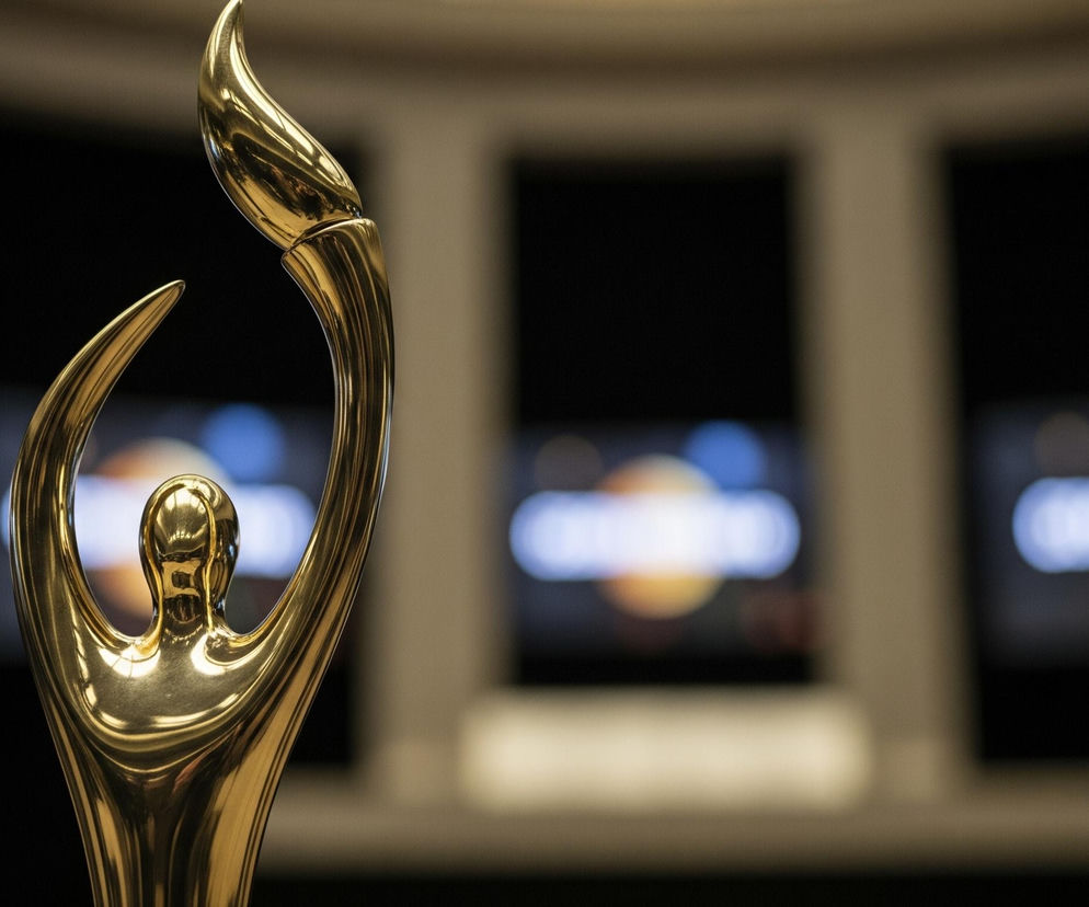 Złota, błyszcząca statuetka o nieregularnym, abstrakcyjnym kształcie dominuje prawą i środkową część kadru, skupiając na sobie większość uwagi. Jej powierzchnia odbija światło, tworząc refleksy i cienie, które podkreślają jej trójwymiarowość. Statuetka przypomina stylizowaną postać ludzką z ramionami uniesionymi do góry, trzymającą stylizowany płomień lub pióro. Tło jest rozmazane i ciemne, z kilkoma jasnymi, rozmyślonymi plamami światła w odcieniach pomarańczowego, niebieskiego i białego, co sugeruje obecność ekranów lub innych źródeł światła w oddali.
