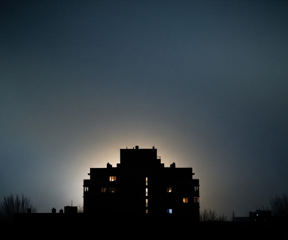 Duży, ciemny budynek mieszkalny o nieregularnej, wielopoziomowej bryle, widoczny jest w centrum kadru, jako ciemna sylwetka. Za nim rozciąga się gradientowe tło, od jasnożółtego światła za budynkiem, przechodzące w błękit i ciemny granat w górnej części. W oknach budynku świecą żółte i jedno niebieskie światło, tworząc kontrastujące, jasne plamy. Na dole, po obu stronach budynku, widoczne są zarysy nagich drzew i innych, mniejszych zabudowań, również w ciemnej sylwetce.