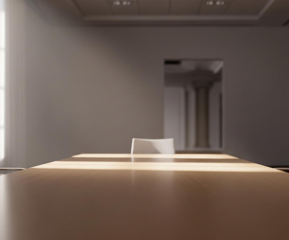 Drewniany stół konferencyjny, o jasnobrązowej barwie, dominuje na pierwszym planie, rozciągając się przez całą szerokość obrazu. Na jego powierzchni widoczne są równoległe pasma intensywnego, jasnożółtego światła słonecznego. Po obu stronach stołu stoją beżowe krzesła z metalowymi, połyskującymi ramami, z czego jedno jest częściowo widoczne po lewej stronie, a drugie po prawej. W tle, po lewej stronie, rozciąga się duże okno z jasnymi, pionowymi i poziomymi szprosami, przez które wpada silne światło. Na ścianie naprzeciwko stołu znajduje się prostokątny otwór prowadzący do innego pomieszczenia, gdzie widać niewyraźne kolumny. Ciemne, drewniane drzwi są widoczne w prawym górnym rogu obrazu.