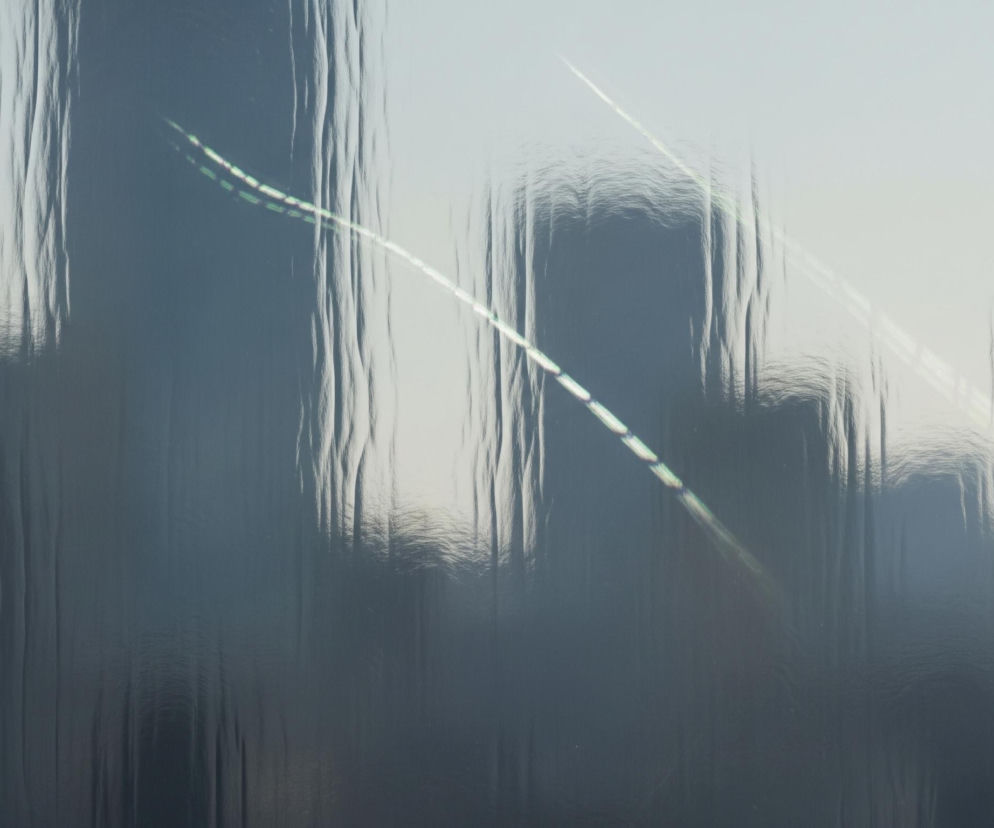 Ciemne, pionowe sylwetki wysokich budynków miejskich, zamazane i zdeformowane przez obecność kropli wody lub innej cieczy na przezroczystej powierzchni. Tło za budynkami jest jasnoszare. Przez zniekształconą perspektywę przebiegają dwie jasne, zygzakowate linie, które wydają się odbiciami światła, posiadając zielonkawe i białe segmenty. Całość obrazu jest rozmyta i nieostra, z widocznymi refleksami światła na falistej strukturze powierzchni.