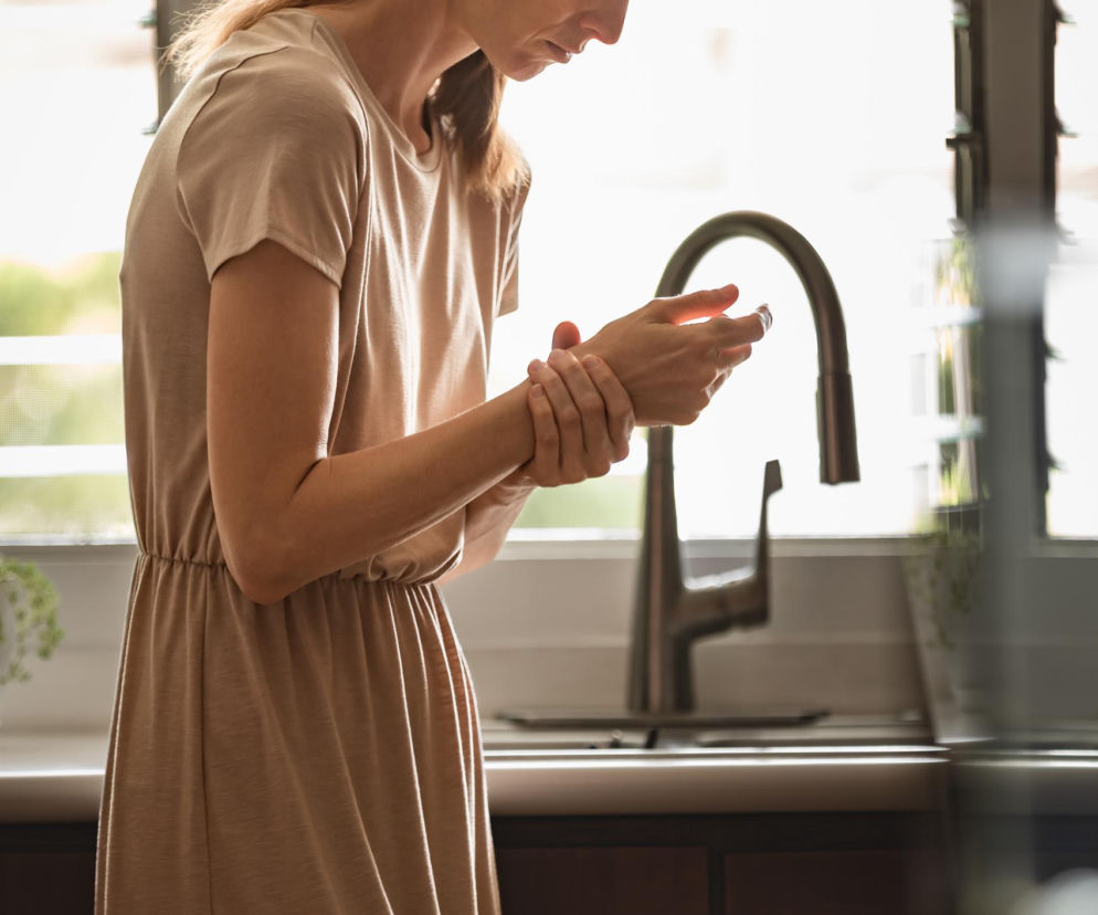 Kobieta w beżowej sukience uciskająca nadgarstek lewej ręki, stojąca w kuchni przy zlewie z baterią i oknem w tle. Scena symbolizuje ból nadgarstka przy krojeniu, o którym można przeczytać więcej na Poradnik Zdrowie.