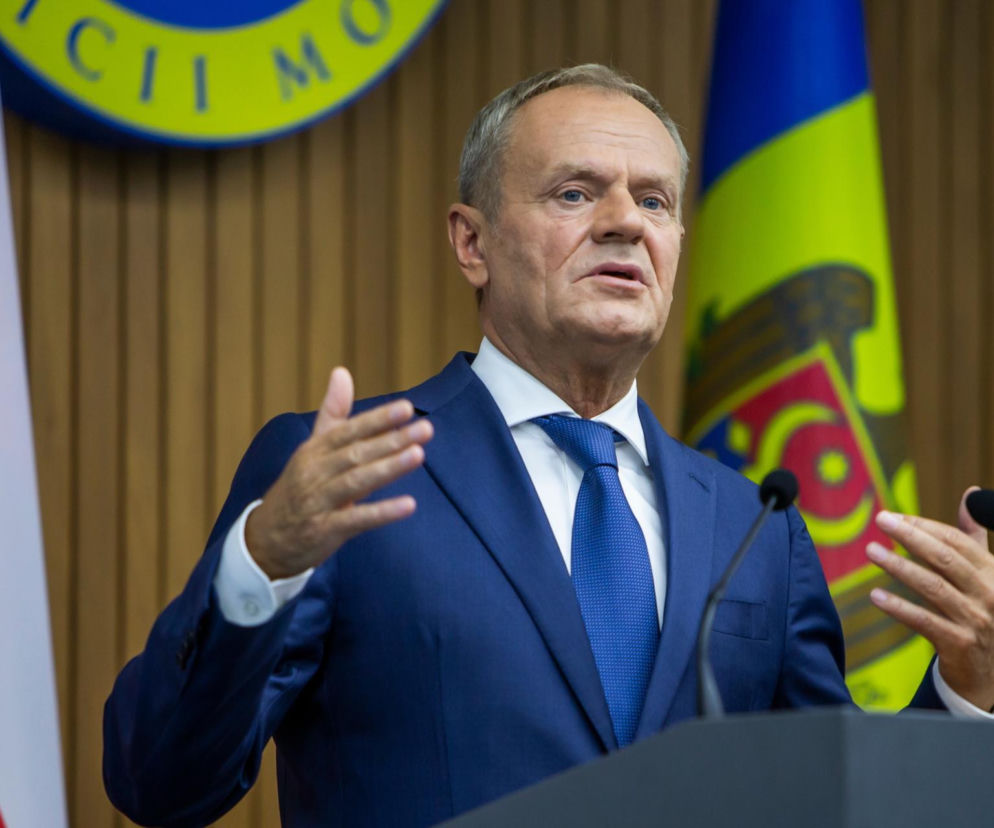 Premier Donald Tusk w granatowym garniturze i krawacie, gestykulujący podczas wystąpienia publicznego, na tle flag Polski i Mołdawii. Dyskusje o proteście wobec KSeF z fakturą na premiera przeczytasz na Super Biznes.