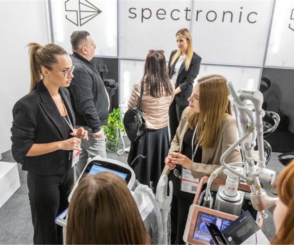 Kobiety i mężczyźni na stoisku wystawienniczym firmy Spectronic podczas targów Estetica Expo, prezentujący urządzenia do medycyny estetycznej i kosmetologii. Dowiedz się więcej o nowościach branżowych na Poradnik Zdrowie.