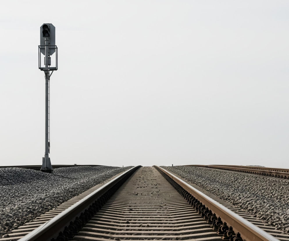 Centralnym punktem kompozycji jest wysoki, szary semafor kolejowy, umieszczony po lewej stronie kadru, ze swoimi ciemnymi soczewkami widocznymi na tle jasnego, bezchmurnego nieba. Pod semaforem biegną dwa tory kolejowe, rozchodzące się symetrycznie w stronę horyzontu i zbiegające w oddali, ułożone na szarym tłuczeniu kamiennym, które pokrywa większą część dolnej połowy zdjęcia. Podkłady kolejowe są jasne, a metalowe szyny mają lekko rdzawy odcień.