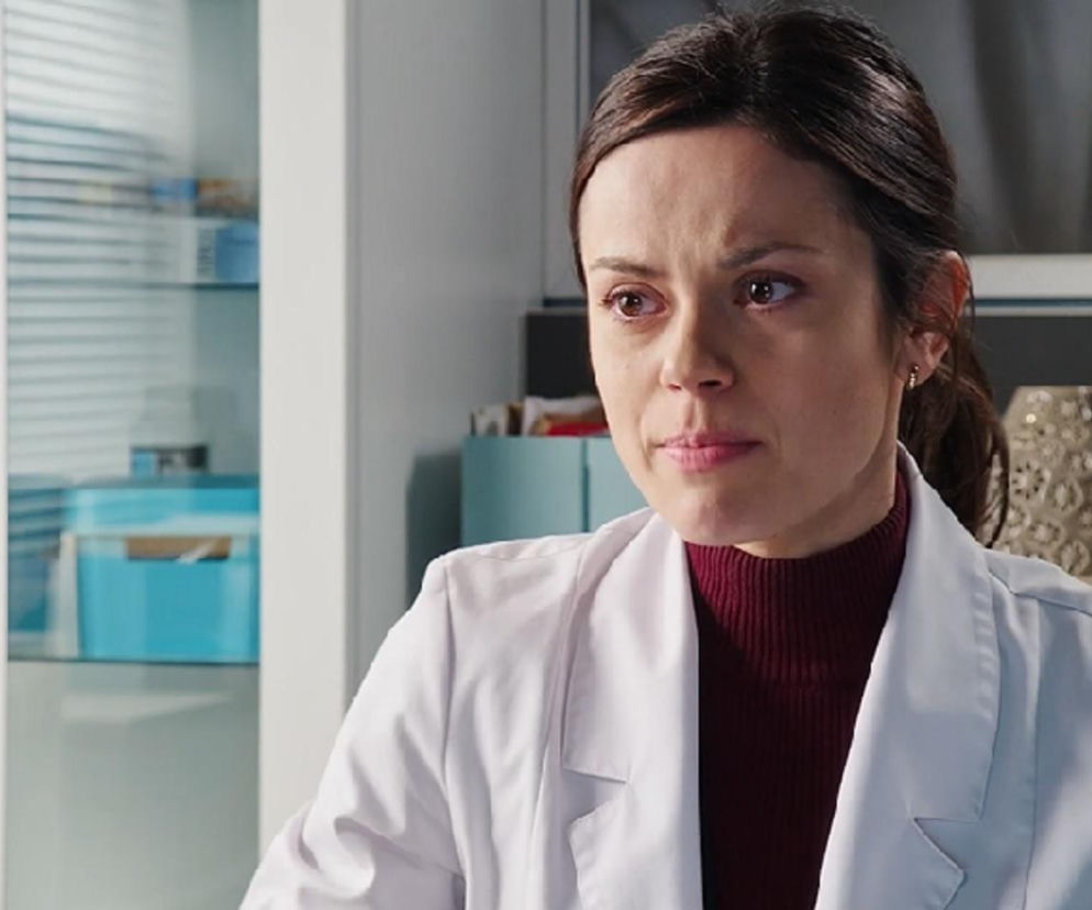 Kasia (Paulina Lasota) z serialu M jak miłość w lekarskim kitlu, z zamyśloną miną, co odzwierciedla jej dylemat związany z porzuceniem pracy w klinice na żądanie Mariusza. O tej i innych perypetiach bohaterów przeczytasz na Super Seriale.