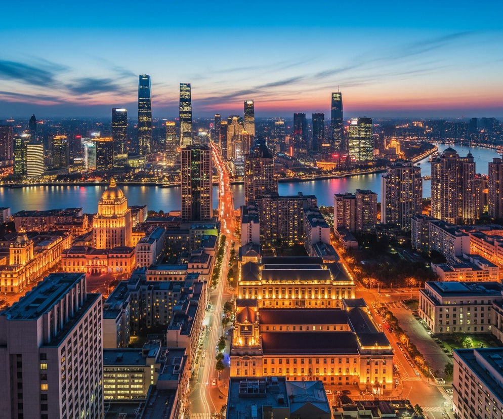 Panorama miasta o zmierzchu ukazuje rzekę biegnącą przez środek, oddzielającą dwa brzegi zabudowane gęsto oświetlonymi budynkami. W tle, na horyzoncie, dominują liczne, wysokie wieżowce z rozświetlonymi oknami, niektóre z nich wyróżniają się smukłą, nowoczesną architekturą. Na pierwszym planie, po lewej stronie, widoczne są klasyczne, monumentalne budynki o jasnej, ciepłej elewacji, intensywnie podświetlone, co kontrastuje z ciemniejącym niebem w odcieniach błękitu i różu. Przez centrum kadru biegnie szeroka ulica, na której smugi świateł pojazdów tworzą długie, pomarańczowe linie.