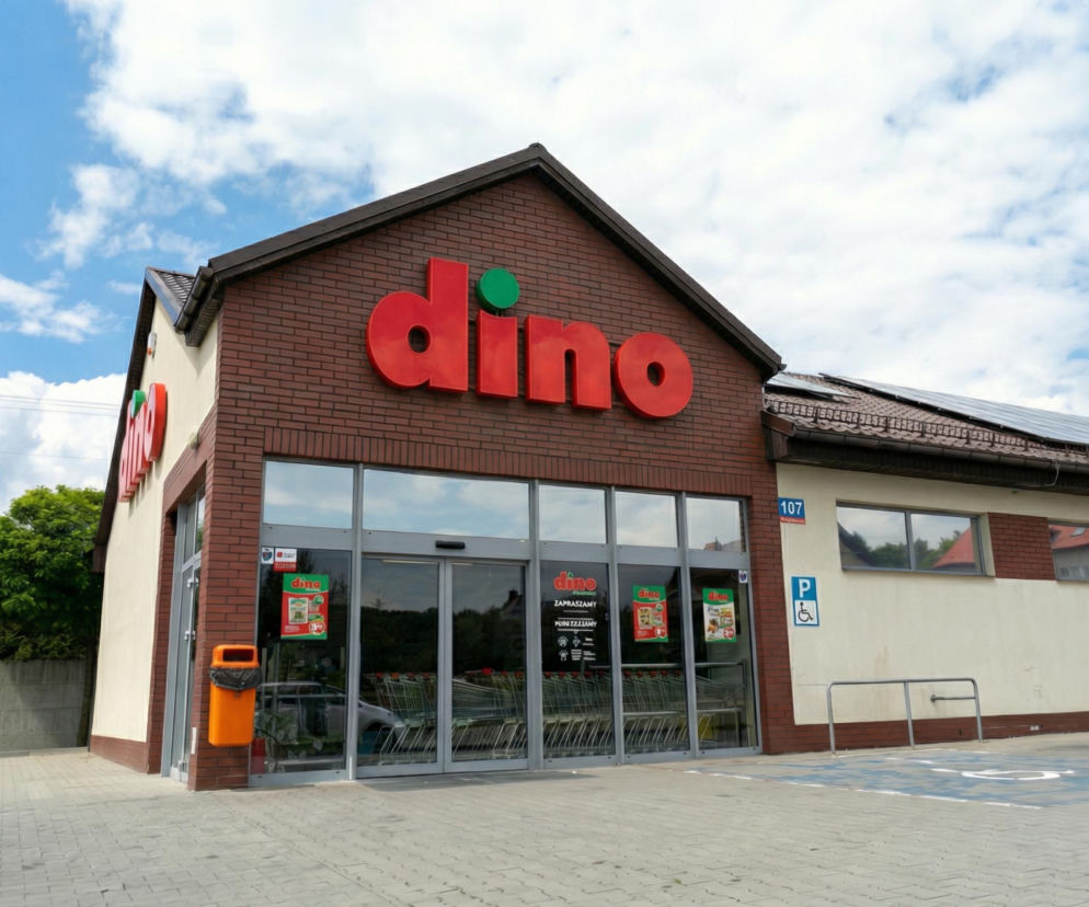 Sklep sieci Dino z czerwonym logo na tle ceglanej fasady i białych ścian. Widoczne szklane drzwi wejściowe z plakatami promocyjnymi oraz wózki sklepowe. Dach budynku posiada panele słoneczne, a przed wejściem znajduje się pomarańczowy kosz na śmieci i niebieskie oznakowanie miejsca parkingowego dla niepełnosprawnych, co jest istotną informacją dla czytelników Super Biznes.