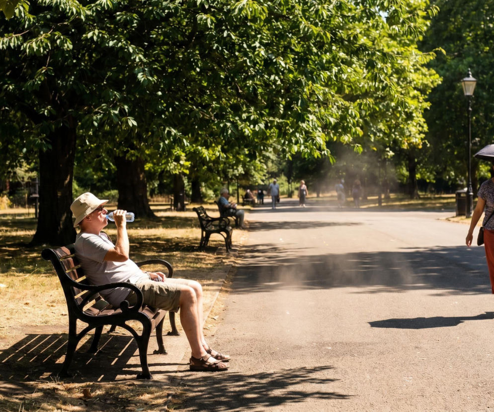 Mężczyzna w kapeluszu pije wodę, siedząc na ławce w parku, a kobieta chroni się pod parasolem, idąc alejką w upalny dzień. Scena obrazuje groźne skutki fali upałów, o których przeczytasz więcej na naszym portalu.