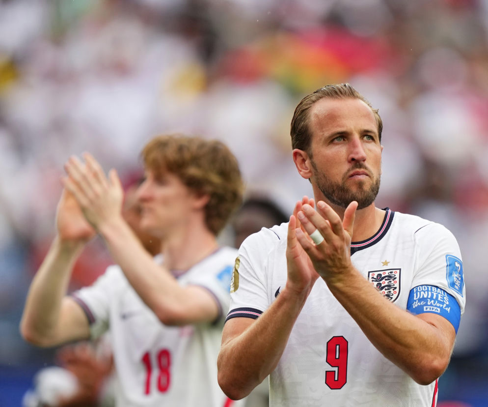 Piłkarz Harry Kane w białej koszulce reprezentacji Anglii z numerem 9 na piersi i opaską Football unites the world FIFA na ramieniu, składa dłonie w geście aplauzu. W tle, częściowo niewyraźna, inna postać o jasnych, kręconych włosach również klaszcze. Na naszym portalu znajdziesz informacje o meczach Anglii.