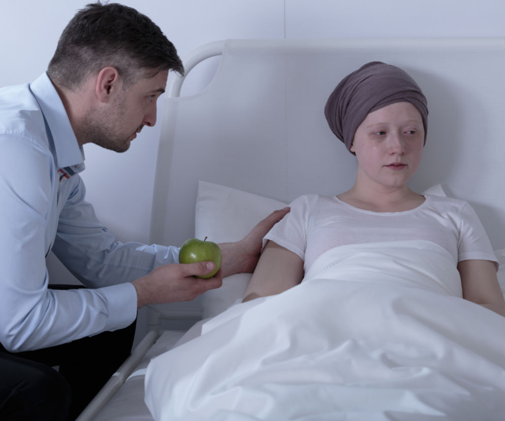 Mężczyzna w błękitnej koszuli podaje zielone jabłko chorej kobiecie w chuście na głowie, leżącej w szpitalnym łóżku, wspierając ją podczas chemioterapii. Więcej o łagodzeniu mdłości przeczytasz na Poradnik Zdrowie.