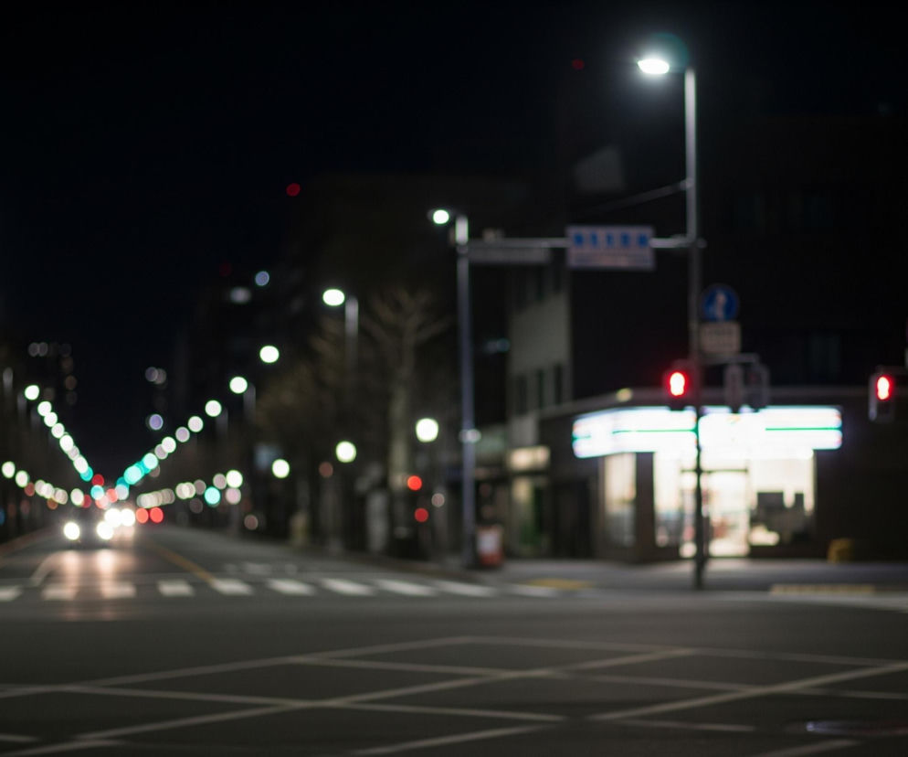Ulica miasta nocą, widziana z poziomu drogi, z licznymi rozmyciami świateł. W centrum obrazu znajduje się rozległe skrzyżowanie z białymi liniami pasów i przejść dla pieszych, które odbijają światło. Po obu stronach drogi widoczne są latarnie uliczne z jasnymi, białymi światłami, tworzące świetlisty ciąg, który zbiega się w oddali. Na pierwszym planie po prawej stronie widać czerwone światło sygnalizacji ulicznej, obok którego znajduje się jaśniej oświetlony budynek z zielono-białym neonowym szyldem, prawdopodobnie sklep. W tle widać smugi świateł samochodów nadjeżdżających z naprzeciwka, a także dalsze latarnie i niewyraźne zarysy budynków.