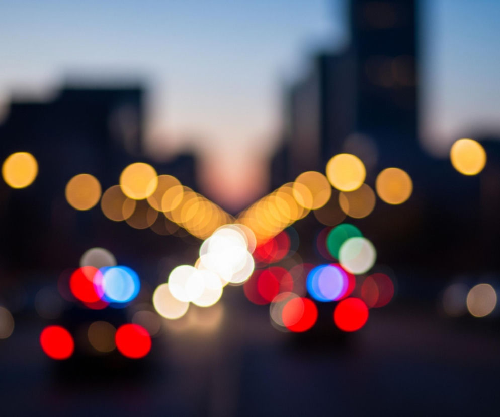 Rozmyty obraz przedstawia nocną lub wieczorną scenę miejską, gdzie dominują liczne, okrągłe światła bokeh. W centralnej części kadru widoczny jest strumień jasnych, żółtych i białych świateł, biegnących w głąb obrazu, sugerując ulicę. Po obu stronach tego strumienia znajdują się czerwone i niebieskie światła, przypominające światła tylne i sygnałowe samochodów. Tło stanowią ciemne, rozmyte kształty budynków oraz niebiesko-fioletowe, przechodzące w błękit niebo.