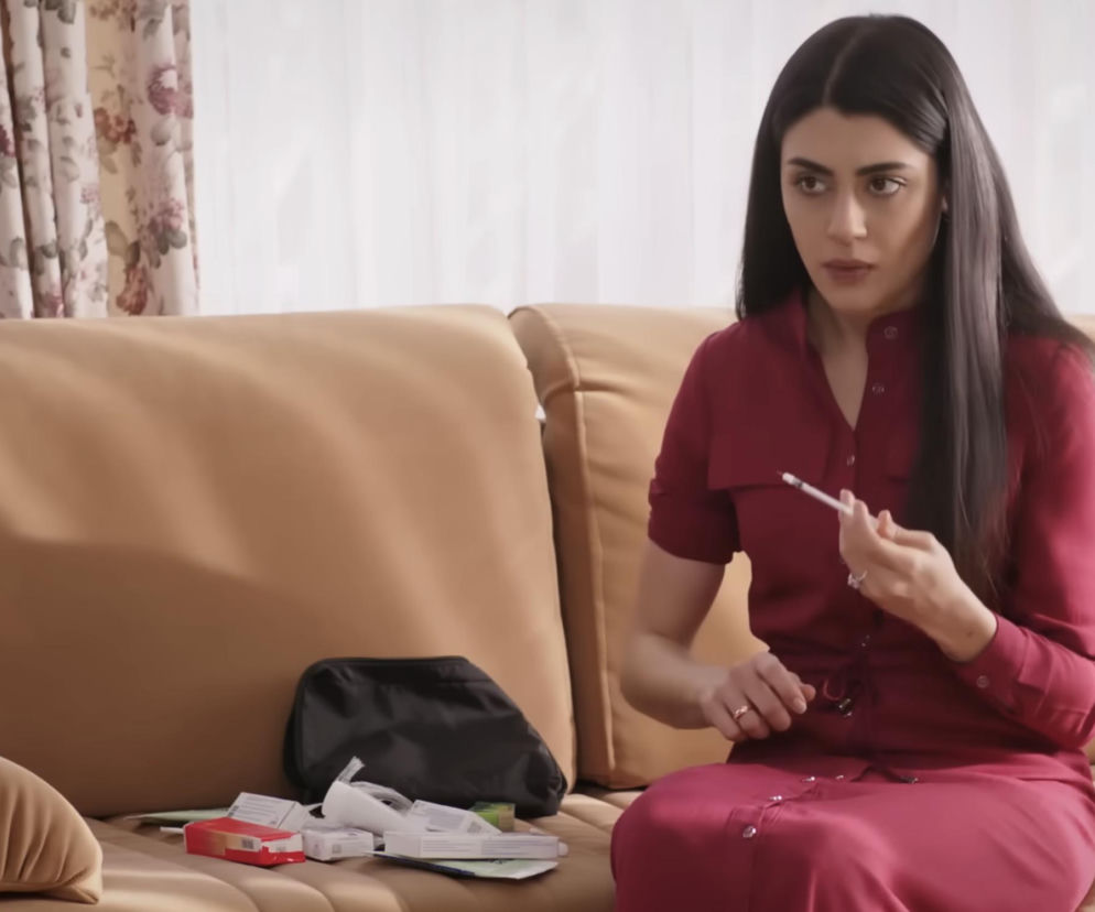 Młoda kobieta, Hancer, w bordowej sukience siedzi na beżowej kanapie, trzymając strzykawkę z niepokojem. Na kanapie obok niej leżą pudełka z lekami i czarna kosmetyczka. Scena z serialu Panna młoda, gdzie Hancer ma być oszukana i być może bezpłodna. Więcej o intrygach w Super Seriale.