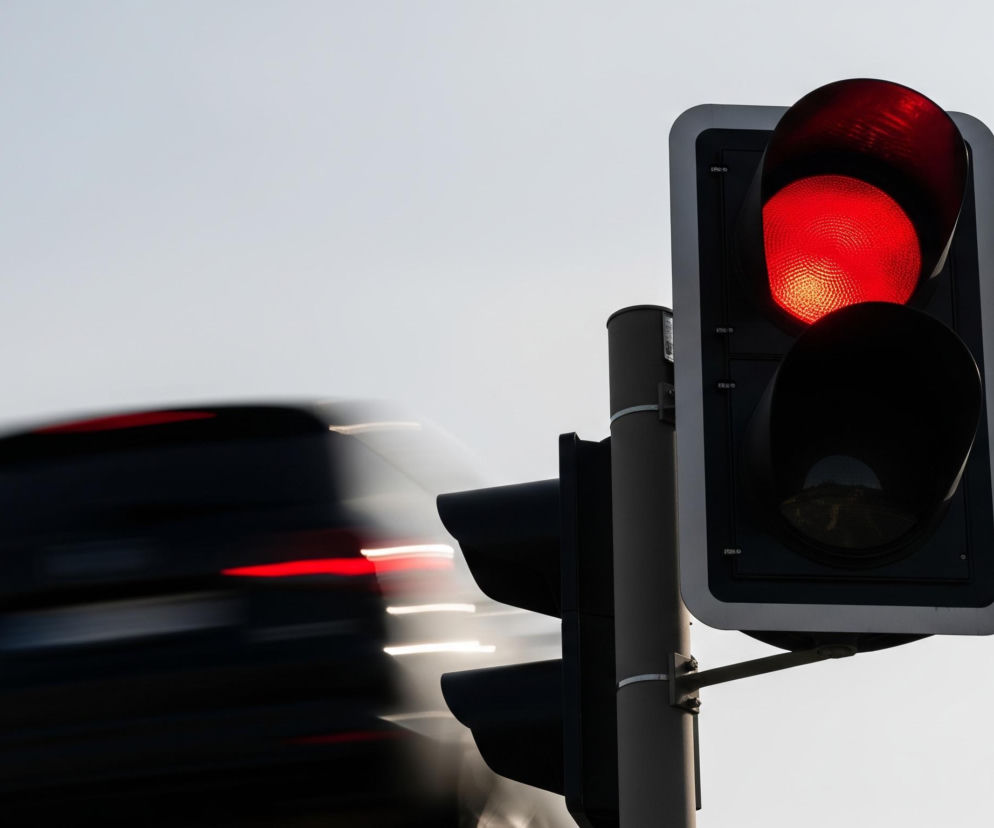 Głównym obiektem jest pionowa sygnalizacja świetlna z czarną obudową i wyraźnie świecącym czerwonym światłem u góry, o widocznej, okrągłej, teksturowanej powierzchni. Sygnalizacja jest zamontowana na szarym słupie, widocznym w prawym centrum kadru, a poniżej czerwonego światła znajdują się dwa kolejne, zgaszone, czarne klosze. Za sygnalizacją, w lewej części obrazu, rozmyty jest ciemny samochód z jaskrawymi czerwonymi i białymi smugami świateł, co sugeruje jego ruch. Tło stanowi jednolite, jasnoszare, niemal białe niebo.