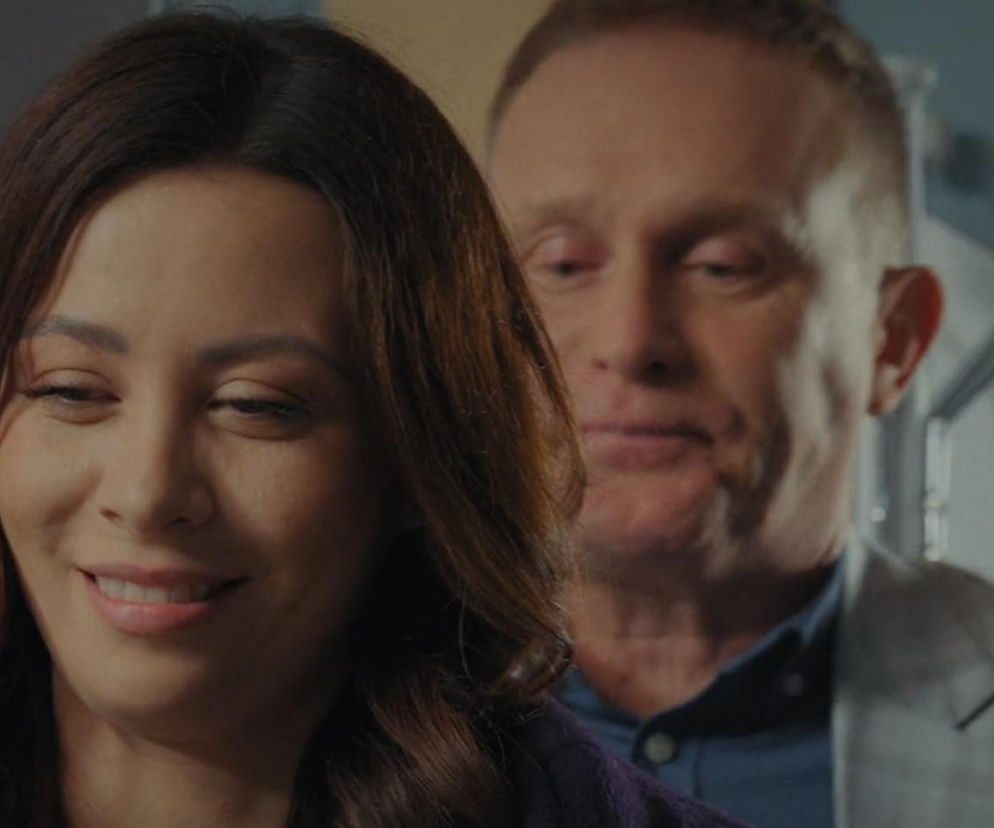 Kobieta o brązowych włosach, z delikatnym uśmiechem na twarzy, patrzy w dół, a w tle widać lekko rozmazaną postać mężczyzny. Zdjęcie przedstawia Joannę i Artura z serialu M jak miłość. Artykuł o ich wspólnych planach przeczytasz na portalu Super Seriale.