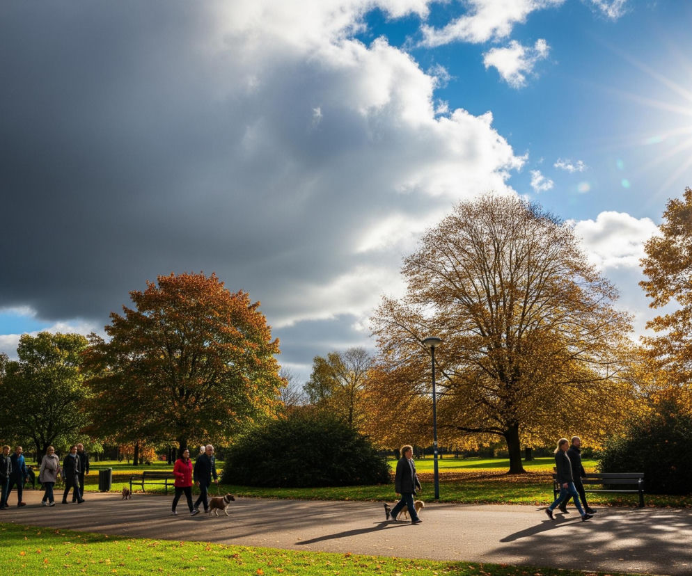 Grupa ludzi spaceruje po parku w słoneczny jesienny dzień, idąc ścieżką wijącą się przez teren zielony pokryty opadłymi liśćmi. Po lewej stronie górę nieba zajmuje duża, ciemna, ciężka chmura burzowa, podczas gdy po prawej stronie dominuje jasne, słoneczne niebo z rozproszonymi białymi chmurami. Drzewa o pomarańczowo-żółtych liściach jesiennych są rozrzucone po całym parku, oświetlone promieniami słońca wpadającymi z prawej górnej części kadru.
