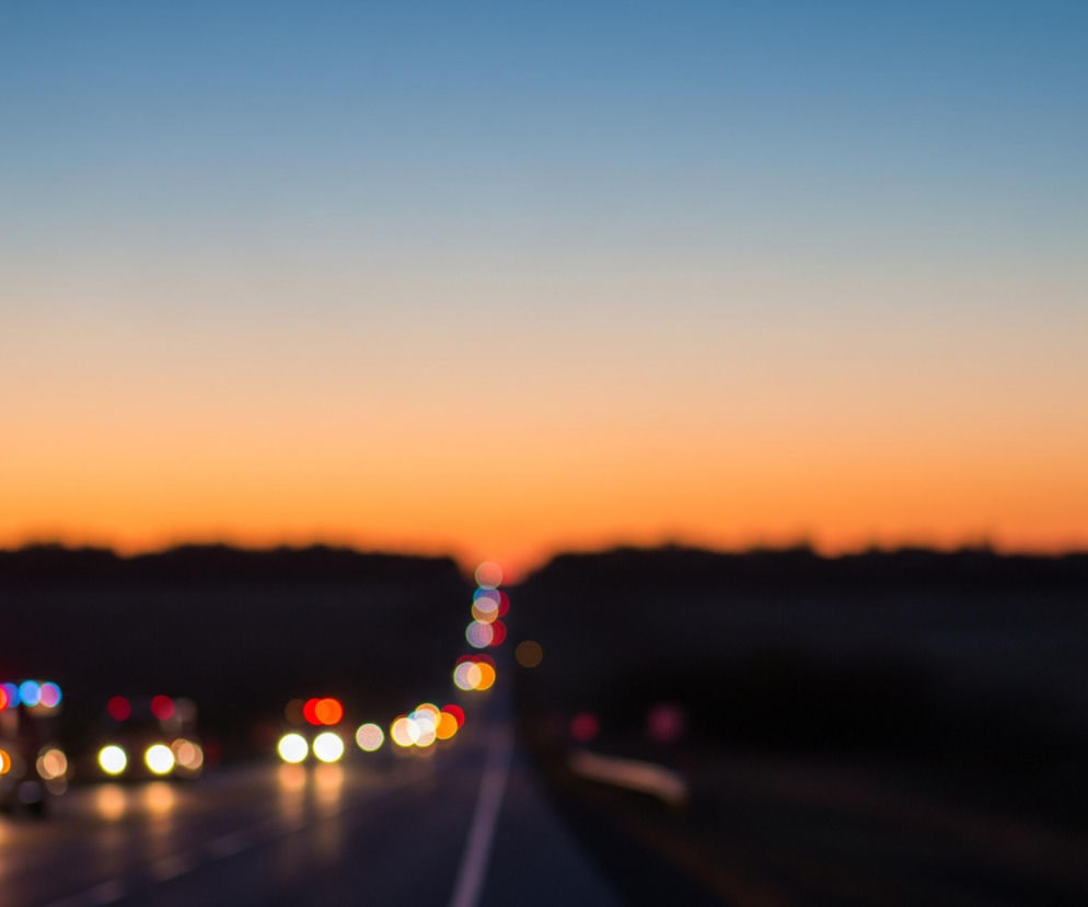 W centrum kadru rozciąga się droga szybkiego ruchu w kierunku horyzontu, z niewyraźnymi światłami samochodów tworzącymi kolorowe, okrągłe plamy. Na pierwszym planie po lewej stronie znajdują się rozmyte światła pojazdu, prawdopodobnie ambulansu, z wyraźnie widocznymi czerwonymi i niebieskimi światłami sygnalizacyjnymi oraz dwoma jasnymi, żółto-białymi reflektorami. Horyzont dominuje gradient nieba, przechodzący od głębokiego błękitu na samej górze, przez jaśniejszy błękit, do ciepłego pomarańczowego i żółtego odcienia u dołu, tuż nad ciemnym, zamazanym pasem lasu lub zarośli.