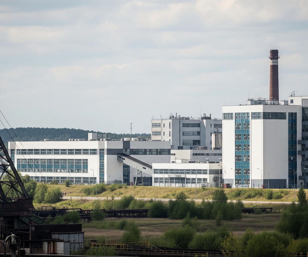Na pierwszym planie, po lewej stronie kadru, dominuje wysoka, metalowa konstrukcja wieży szybowej w ciemnych barwach, częściowo zardzewiała, z widocznymi kołami linowymi na górze. W tle, po prawej stronie, rozciąga się kompleks nowoczesnych, biało-szarych budynków przemysłowych z licznymi oknami o błękitnych szybach, połączonych pochylniami. Ponad nimi, po prawej stronie, wznosi się wysoki, ceglany komin z białym pierścieniem u góry. Pomiędzy starym i nowym kompleksem oraz na pierwszym planie, znajduje się trawiasty teren z rzadkimi krzewami i kilkoma zarośniętymi torami kolejowymi lub konstrukcjami.