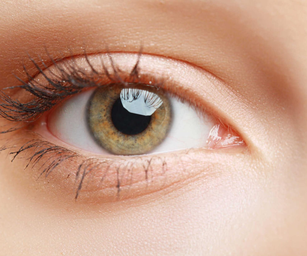 Oko kobiety z widoczną jasnozielono-brązową tęczówką i źrenicą, otoczone długimi, ciemnymi rzęsami, lekko podkreślone makijażem. Skóra wokół oka jest jasna i gładka, a w oku odbija się subtelne światło. Więcej o nietypowych objawach zaćmy przeczytasz na Poradnik Zdrowie.