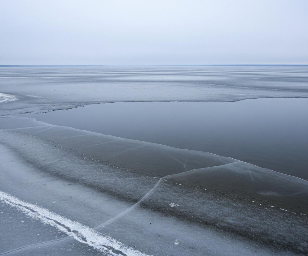 Rozległy, zamarznięty akwen wodny pod pochmurnym, bladoniebieskim niebem. Na pierwszym planie, w lewej dolnej części zdjęcia, widać nieregularne płaty lodu o różnej fakturze, z wyraźnymi pęknięciami i smugami białego śniegu, przypominającymi fale. Część powierzchni lodu jest gładka i ciemniejsza, podczas gdy inne obszary są szorstkie i poprzecinane jasnymi liniami. Na lód padają odcienie szarości i błękitu, z kilkoma drobnymi gałązkami i punkcikami rozrzuconymi na powierzchni. Linia brzegowa, zaznaczona ciemniejszym pasem drzew na horyzoncie po lewej stronie, rozciąga się w oddali. Po prawej stronie, bliżej środka obrazu, otwarta woda w głębokim, spokojnym odcieniu granatowego, płynnie przechodzi w zamarzniętą powierzchnię, tworząc wyraźny kontrast między lodem a lustrem wody.