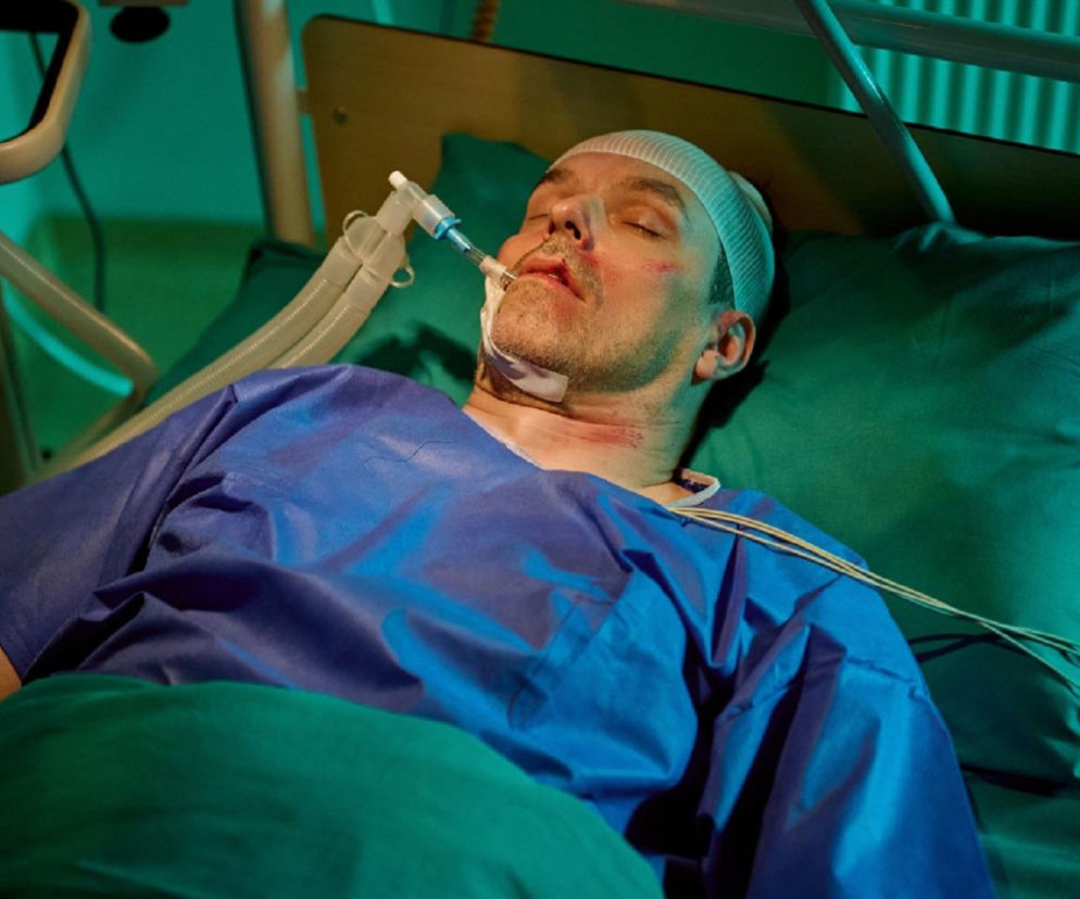 Mężczyzna, Sebastian (Robert Jarociński), leży w szpitalnym łóżku w niebieskiej szpitalnej koszuli, z głową zabandażowaną i aparaturą medyczną podłączoną do ust, ranny po konfrontacji. Na jego twarzy i szyi widać ślady obrażeń. Jego stan jest poważny, walczy o życie, co jest tłem dramatycznych wydarzeń w serialu Na Wspólnej, o czym przeczytasz na Super Seriale.