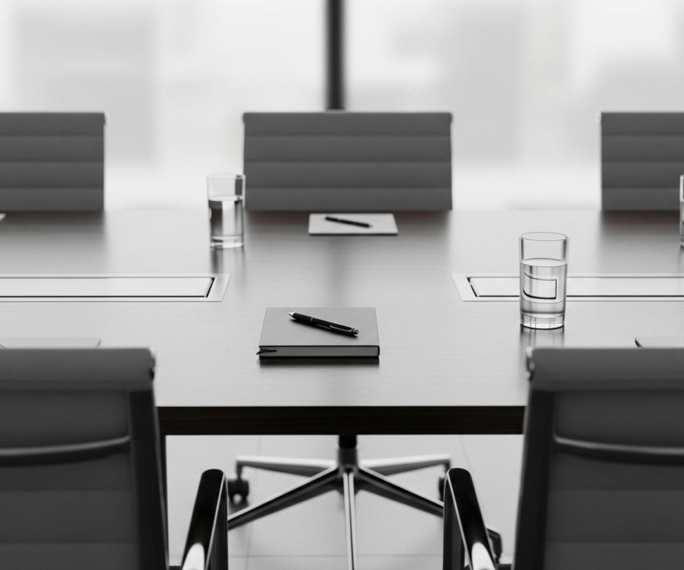 Na pierwszym planie widoczny jest długi, ciemny stół konferencyjny z czarną oprawioną książką i czarnym długopisem leżącym na niej pośrodku. Po obu stronach książki ustawione są szklanki z wodą, a obok nich leżą białe notatniki z czarnymi długopisami. Stół otoczony jest krzesłami z szarymi oparciami. W tle widoczne są zamglone okna, przez które wpada jasne światło.